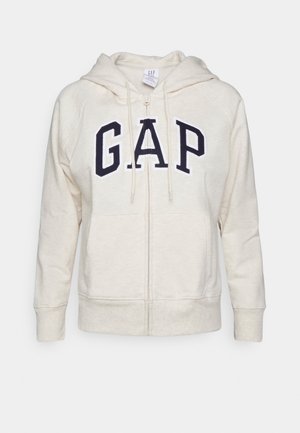 Sweat à capuche crème à fermeture éclair avec capuche à cordon, arborant un grand logo "GAP" bleu marine. Matière douce, poches avant et poignets côtelés.
