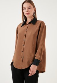 Brown button-up shirt with a black collar, long sleeves, and a relaxed fit. Smooth texture with subtle sheen and side hem slits.
