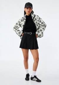 Giacca in pile bianca con motivo floreale nero, dolcevita nero, gonna plissettata nera, cintura nera e sneakers nere con strisce bianche.