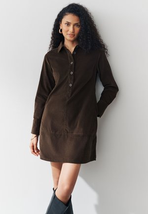 Rjava srajčna obleka iz korduroja z dolgimi rokavi, z gumbi spredaj in stranskimi žepi. Oblečena s črnimi visokimi čevlji. Gladek tekstura, priložnostna zasnova.