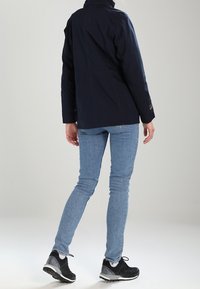 Námořnická modrá voděodolná bunda s vysokým límcem, viditelným šitím a logem, kombinovaná se světle modrými džíny a černými sportovními botami.