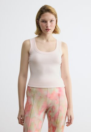 Nuori nainen, jolla on lyhyet vaaleat hiukset, pukeutuneena vaaleanpunaiseen hihattomaan toppiin ja korkeavyötäröisiin housuihin, joissa on vaaleanpunaisia ja beigejä abstrakteja kuvioita.