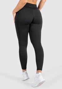 Leggings neri a vita alta realizzati in materiale elastico, con una texture liscia e un design aderente, abbinati a sneakers bianche.