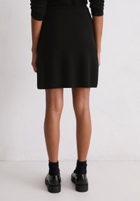 Jupe noire en tricot à coupe droite, avec une large ceinture. Portée avec des chaussettes noires et des chaussures, vue de dos. Texture lisse.