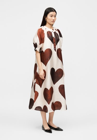 Γυναίκα που φοράει μίντι φόρεμα εκρού με μεγάλα καφέ σχέδια καρδιάς, φουσκωτά μανίκια, κουμπωτό μπροστά και μαύρα μυτερά φλατ, σε πλευρική στάση.