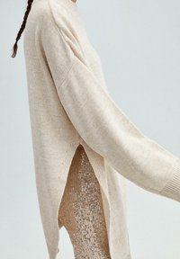 Beige pletený svetr s dlouhými rukávy, který má postranní rozparky odhalující lesklou, flitrovou látku pod ním. Texturovaný, neformální design.