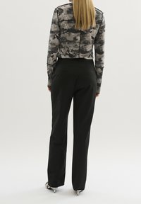 Persona con lunghi capelli biondi che indossa un top a maniche lunghe a strisce nere e grigie, pantaloni neri e tacchi argentati, in piedi davanti a uno sfondo neutro.