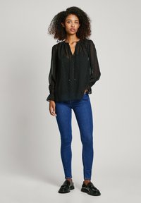 Blusa nera trasparente con scollatura a V, polsini arricciati e dettaglio con fiocco. Indossata con jeans skinny blu e mocassini neri. Tessuto testurizzato con leggera lucentezza.