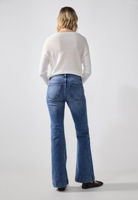 Top de color claro y de manga larga, y jeans acampanados azules. Los jeans cuentan con un diseño clásico de 5 bolsillos y una textura ligeramente desgastada.