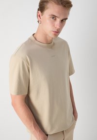 Beige tričko s krátkým rukávem vyrobené z bavlny, s kulatým výstřihem; uprostřed se nachází jemná vyšívaná logo. Hladká textura a volný střih.
