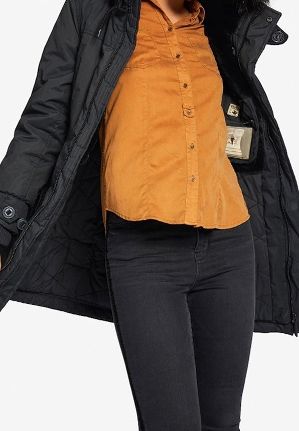 Persona con chaqueta acolchada negra desabrochada sobre una camisa mostaza de botones y pantalones negros de corte ajustado, mostrado desde el cuello hasta las caderas.