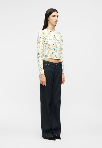 Γυναίκα με μακριά σκούρα μαλλιά, που φοράει λευκό ροζάνεμα με μακριά μανίκια, μαύρο παντελόνι με φαρδιά πόδια και μαύρα μυτερά παπούτσια, στέκεται πλευρικά.