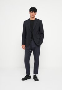 Sacou bleumarin croit cu rever înclinat, tricou negru, pantaloni închiși cu șnur la talie și încălțăminte neagră. Material neted cu o croială structurată.
