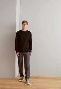 Schwarzes Langarmshirt kombiniert mit grauen Hosen und schwarz-weißen Sneakers; das Model steht auf einem Holzboden vor einem minimalistischem grauen Hintergrund.