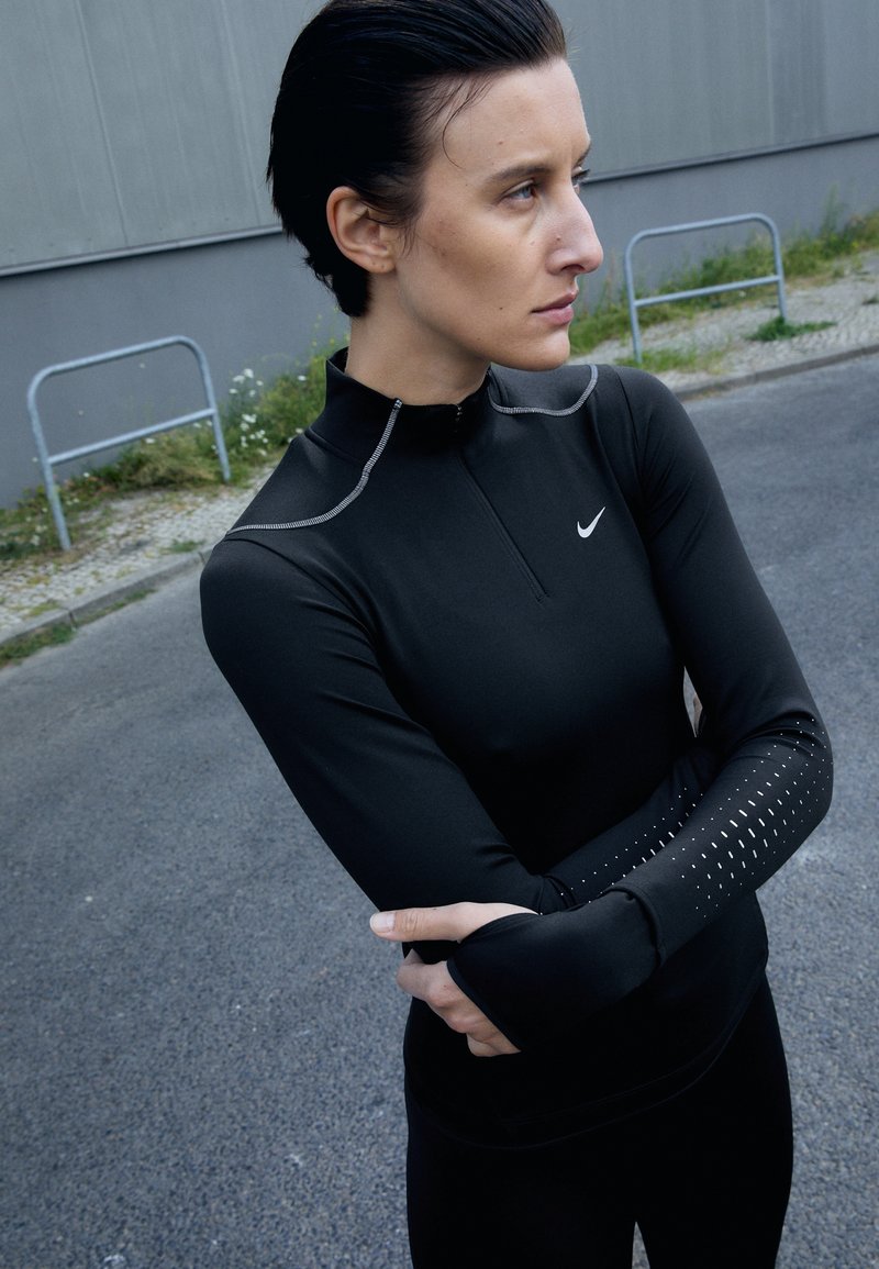 Czarna, długosleeveowa koszulka sportowa Nike z wysokim kołnierzem, przednim zamkiem, odblaskowym przeszyciem i perforowanymi akcentami na rękawach.