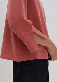 Rust-colored, lightweight sweatshirt with 3/4 sleeves and a straight hem. Soft fabric and minimal stitching detail, paired with dark denim.