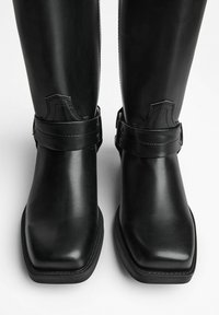 Black leather knee-high boots with a square toe, featuring buckle accents and contrasting stitching around the ankle strap.