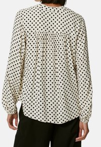 Hvid bluse med sorte prikker, lange ærmer med elastiske cuffs og rynket detalje på ryggen. Glat stof med en løs pasform.