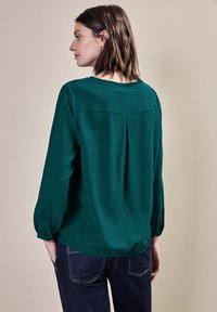 Tealblus med långa, samlade ärmar och en strukturerad yta. Har en plisserad rygg och en avslappnad passform. Bärs med mörk denim.
