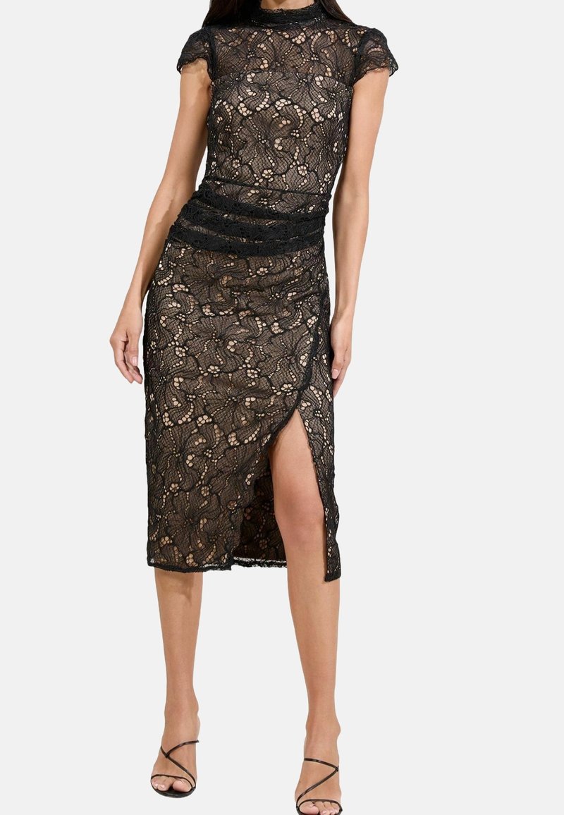 Femme portant une robe midi en dentelle noire avec manches courtes et une fente à l'avant, associée à des sandales noires à talons hauts à brides.