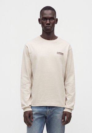 Mann trägt beige Langarmshirt mit kleinem schwarzem Text auf der Brust und blaue Jeans, steht vor weißem Hintergrund.