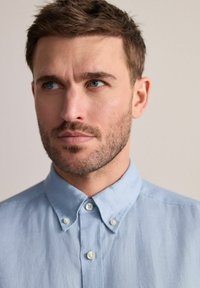 Camisa de botones azul claro con cuello y dos botones en el cuello. La tela parece suave y ligeramente texturizada.