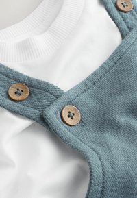 Prim-plan cu o cămașă albă cu dungi sub salopeta albastră texturată, având doi nasturi maro din lemn.