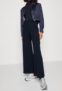 Mörkblå kort satinjacket, svart polotröja, vida mörkblå byxor och silvriga sneakers. Slät textur, elegant design, modern atletisk stil.