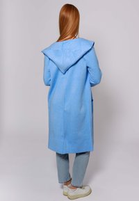 Cappotto lungo azzurro chiaro con cappuccio, dal tessuto morbido, con cucitura centrale e tasche, indossato sopra jeans in denim chiaro e sneakers bianche.