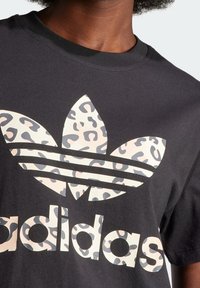 Närbild av en person som bär en svart t-shirt med en stor Adidas-logotyp i en beige och grå leopardmönster.