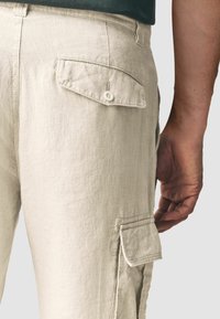 Pantalones cargo color beige claro, confeccionados en tela texturizada, con un bolsillo trasero abotonado y bolsillos laterales tipo parche. Primer plano de la cinturilla y la pierna.