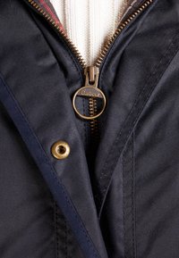 Detail blízko zipu tmavě modré bundy s mosazným kroužkem na tahání Barbour, drukem a bílým pleteným svetrem viditelným pod ním.