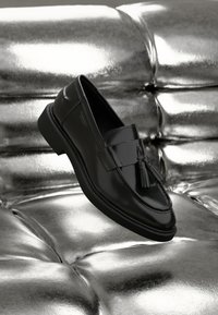 Black leather loafers with a flat sole, featuring a decorative tassel and stitched detailing, resting on a metallic, cushioned surface.