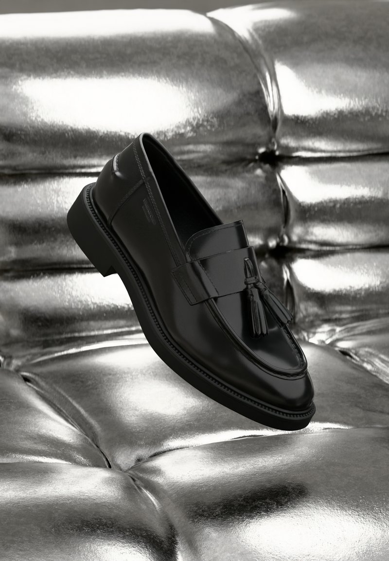Mustad mustad nahast loaferid, millel on tasane tall ja dekoratiivne tuti ning õmbluste detailid, asetatud metallilisest, pehmest pinnast.