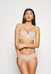 Beige bra with smooth, molded cups and thin adjustable straps. Matching beige brief with subtle logo detail. Minimalist design and seamless texture.