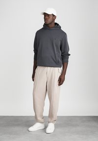 Grå hettegenser, beige vide bukser og hvite joggesko. Hettegenseren har et teksturert stoff og kengurulomme. Enkel hvit caps.