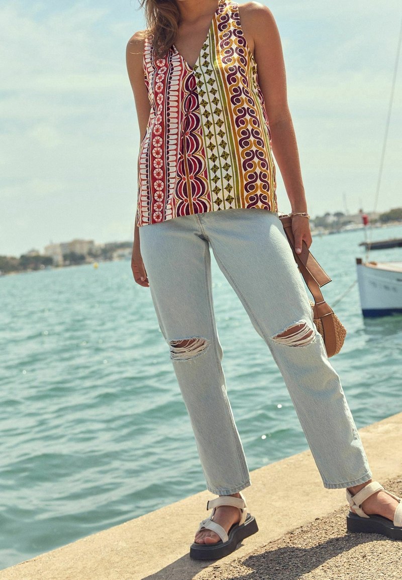 Femme debout au bord de l'eau, portant un haut sans manches à motifs colorés, un jean bleu clair déchiré et des sandales compensées blanches, tenant un sac beige.