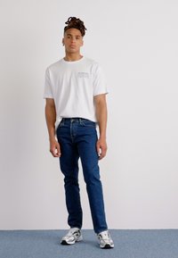 Hvit bomulls-t-skjorte med trykt tekst på brystet, sammen med blå denimjeans. Modellen har på seg grå og hvite joggesko. Enkelt bakgrunn.