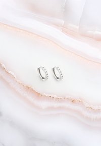 Orecchini a cerchio in argento con una fila di piccole gemme trasparenti lungo un bordo, posizionati su uno sfondo di pietra liscia e di colore chiaro.