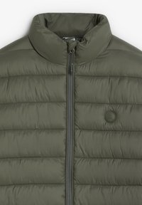 Chaqueta acolchada verde con cuello alto, cierre de cremallera y paneles cosidos. Presenta un logotipo redondo en tono sobre tono en el lado izquierdo del pecho.