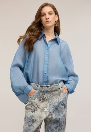 Kvinde med langt bølget hår iført oversized lyseblå bluse, bukser med blomsterprint, blåt bælte med guldlås og hængende øreringe.