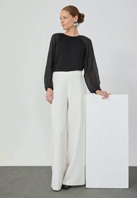 Schwarze Bluse mit langen Ärmeln und gerafft verarbeitetem Ausschnitt, kombiniert mit hoch geschnittenen weißen weiten Hosen, vor einem neutralen Hintergrund.