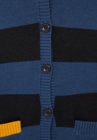 Blå och svart randig kofta med ribbad textur, knappar fram, och en gul accentficka längst ner till vänster.