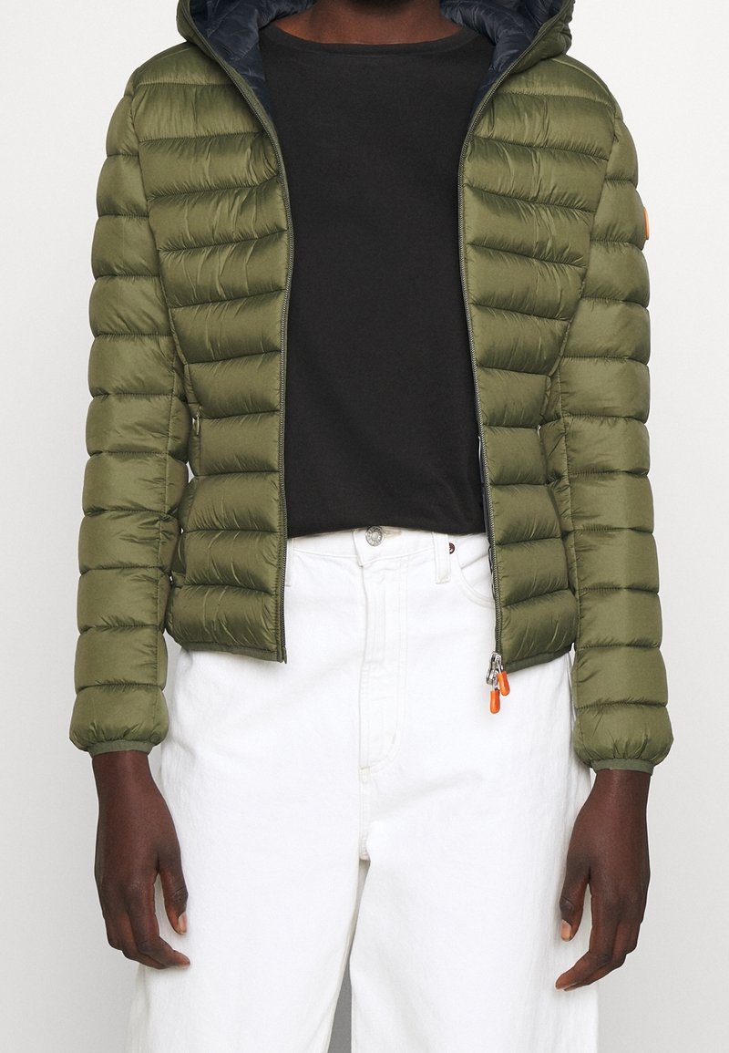 Personne portant une doudoune vert olive ouverte par-dessus un t-shirt noir et un pantalon blanc taille haute sur un fond uni.