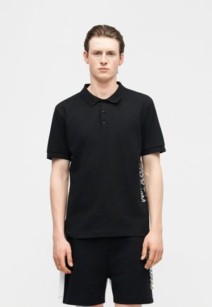 Jeune homme à la peau claire et aux cheveux coiffés en arrière, portant un polo noir à manches courtes et un short noir avec des bandes graphiques blanches sur les côtés.