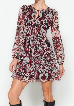 Femme portant une mini-robe à manches longues avec un motif abstrait rouge, noir et blanc et un petit détail d'encolure en trou de serrure à l'avant.