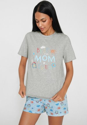 Frau mit langen schwarzen Haaren, die ein graues T-Shirt mit dem Text "Best Mom Ever" und hellblaue Shorts mit Mustern aus Sonnen, Wolken und Häusern trägt.