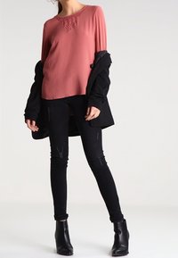 Langärmliges, rosafarbenes Oberteil mit strukturiertem Ausschnitt, kombiniert mit schwarzen Skinny-Jeans und schwarzen Knöchelstiefeln, vor einem neutralen Hintergrund.