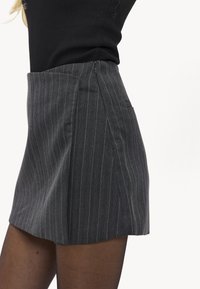 Primer plano de una persona que lleva una falda de talle alto gris oscuro con rayas finas, tableada, y medias negras transparentes sobre un fondo blanco.