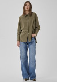 Frau steht mit einer Hand in der Tasche, trägt ein olivgrünes, geknöpftes Langarmhemd, blaue weite Jeans und spitze braune Schuhe.